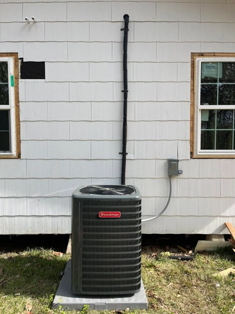 Goodman condenser unit on pad outside a new construction home with vinyl siding and unfinished window framing in West Monroe