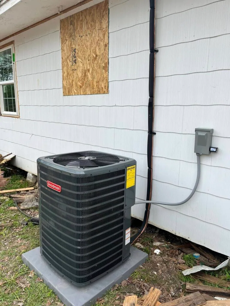 Goodman condenser unit on concrete pad beside white vinyl siding home with plywood patch and electrical disconnect box