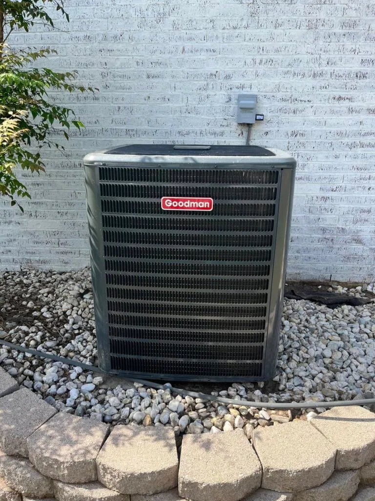 Goodman air conditioner condenser unit on gravel bed against white painted brick wall with stone border
