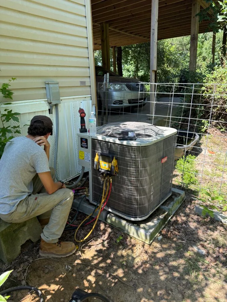 Technician kneeling beside outdoor air conditioner condenser unit connected to refrigerant gauges outside a home in West