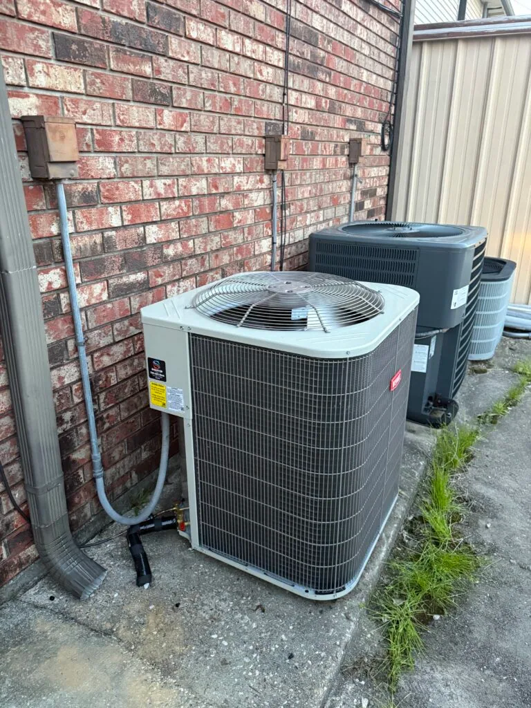 Multiple outdoor air conditioning condenser units lined up against a brick wall behind a commercial building