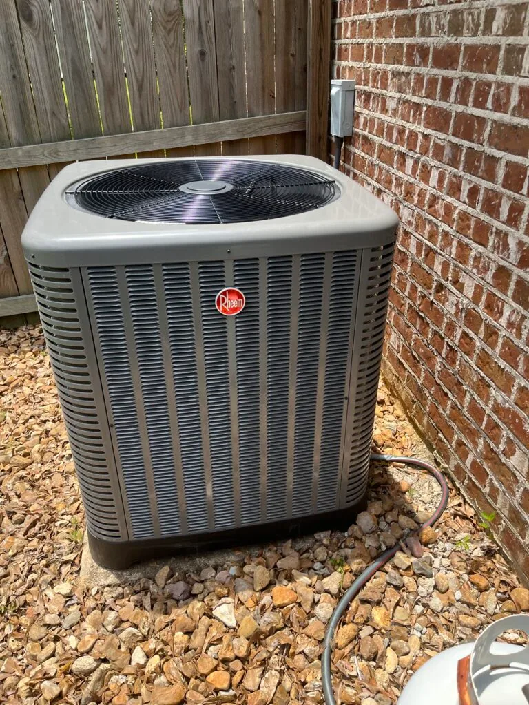 Rheem outdoor condenser unit on gravel bed against a brick wall with electrical disconnect box, wood fence in background