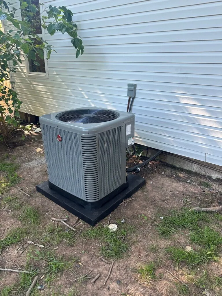 Rheem outdoor condenser unit on a black composite pad beside a mobile home in West Monroe