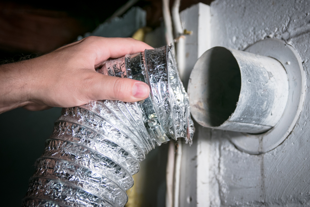 Hand pulling a lint-clogged aluminum flex dryer vent hose off a metal wall vent