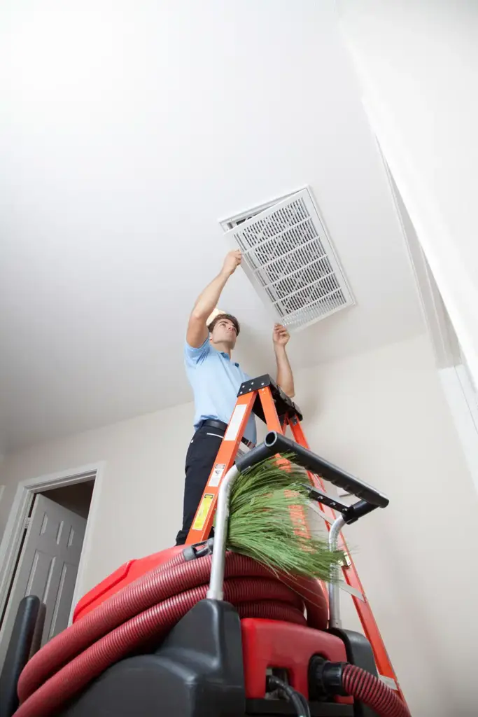 Technician on an orange ladder removing a ceiling return grille above duct cleaning equipment
