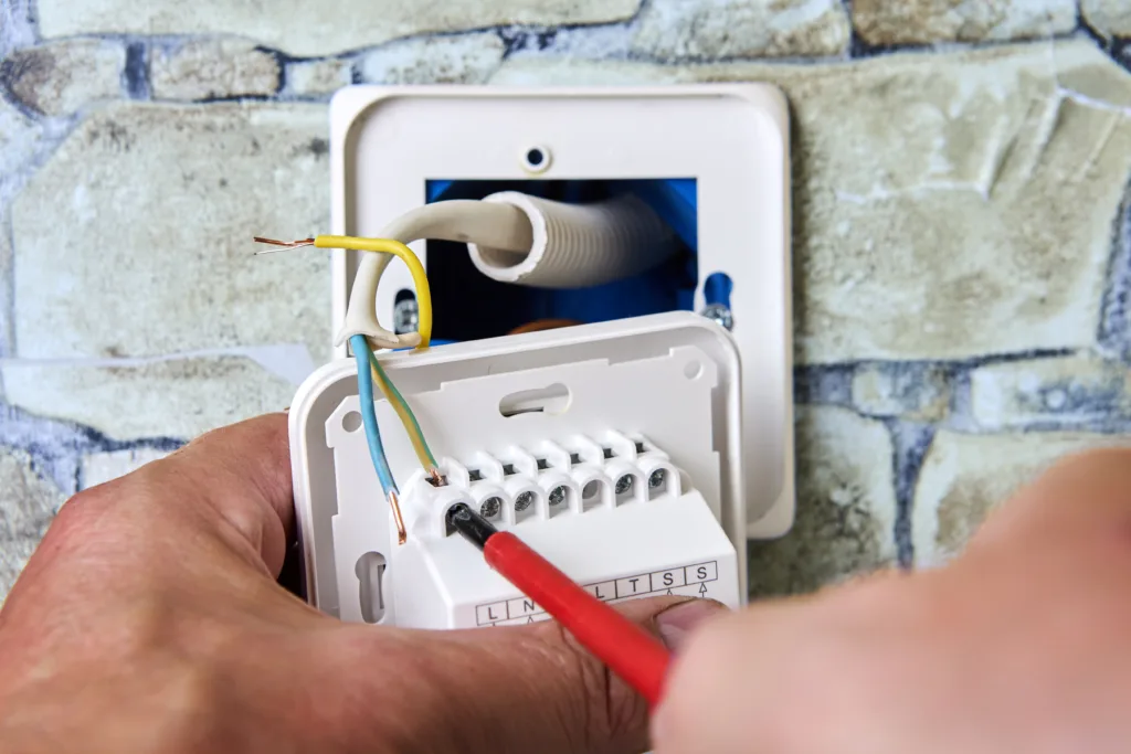 Hands connecting colored wires to a thermostat terminal block with a red screwdriver